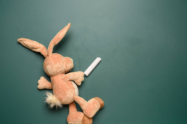 toy bunny holding a piece of chalk on the background of a green chalk school blackboard, place to write - Photo, Image