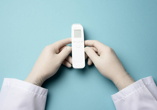 mano en guantes de látex blanco sostienen un termómetro electrónico para medir la temperatura, dispositivo sin contacto - Foto, imagen