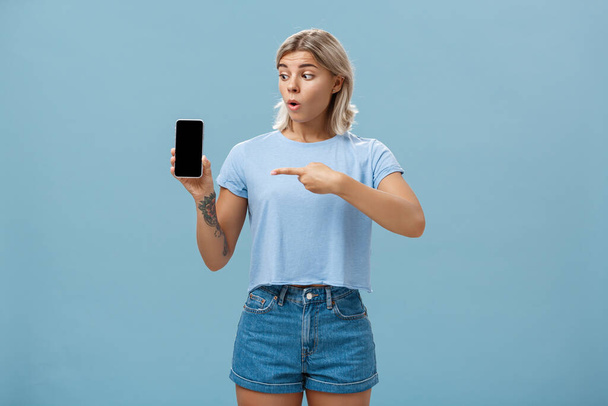 You should download app. Portrait of amazed shocked attractive blonde girl in trendy summer t-shirt holding smartphone gasping from surprise pointing at gadget screen posing over blue background - Foto, afbeelding