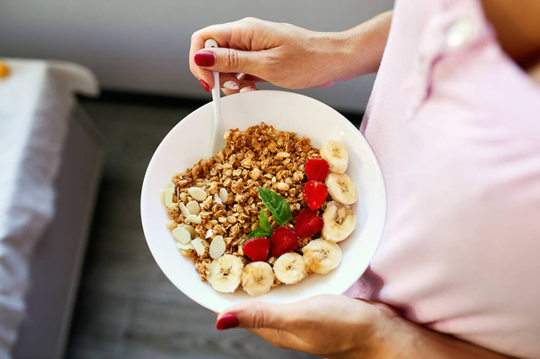 Woman eating healthy breakfast bowl, hold in hand granola, seeds, fresh strawberry, banana, top view, copy space. Clean eating, detox, dieting, vegetarian food concept - Photo, Image