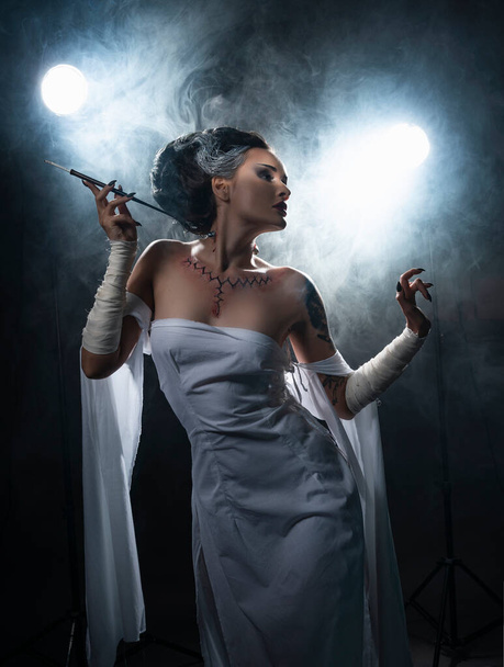 Beautiful girl wearing a Halloween costume with rich make-up and an imitation of an autopsy seam on her chest is emotionally posing holding a cigarette holder in the smoke. Vintage, cosplay design. - Fotoğraf, Görsel