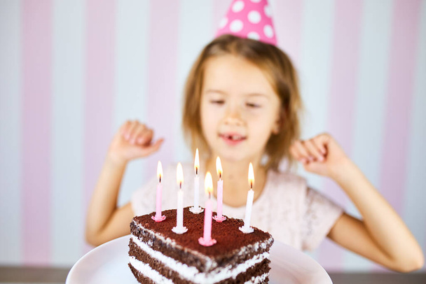 Pieni blondi tyttö hymyilee syntymäpäivä vaaleanpunainen lakki, suklaa syntymäpäivä kakku kynttilöitä. Lapsi juhlii syntymäpäiväänsä. Hyvää syntymäpäivää.. - Valokuva, kuva