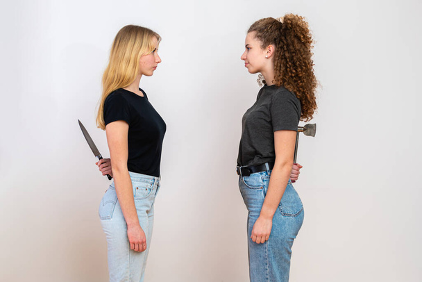 two angry women standing face to face and hiding knife and axe behind back, isolated on white background, the concept of hidden hatred and hypocrisy - Fotó, kép