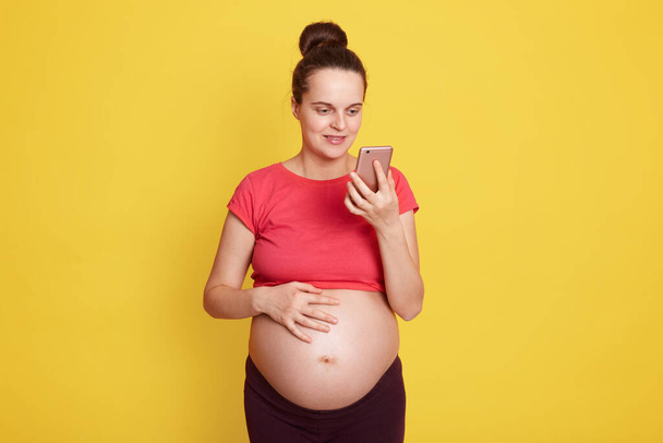 Cheerful pregnant woman using mobile phone chatting with somebody, touching her bare belly, wearing casual t shirt, has hair bun, posing isolate over yellow background. - Valokuva, kuva