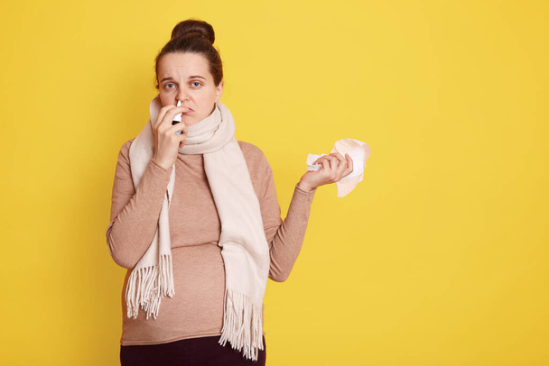 Catching cold during pregnancy, future mom with hair bun dresses beige sweater and scarf, using nasal spray and napkin, posing isolate dover yellow background. - Foto, Imagem