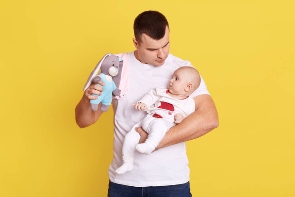 Happy father playing with his baby son or girl with bright rattle, dad holding infant kid and soft toy rabbit in hands, handsome daddy playing with child against yellow wall. - Foto, imagen