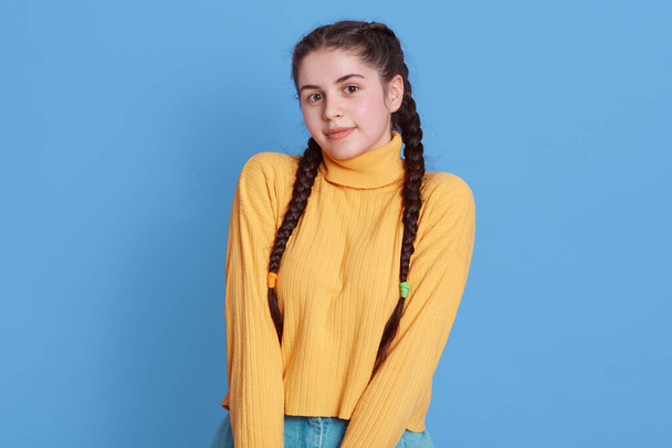 Photo of shy girl with braids in casual outfit, stands smiling isolated on vivid blue color background, wearing yellow casual sweater, looking at camera. - Valokuva, kuva