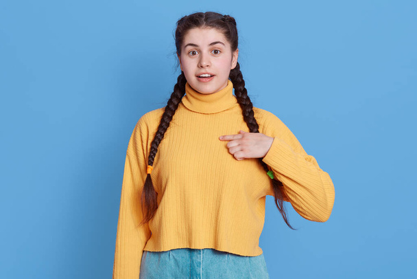 Glad surprised woman points at herself with fingers, cannot believe, being chosen for something, opens eyes widely, wears yellow jumper and jeans, posing isolated over blue background. - Fotografie, Obrázek