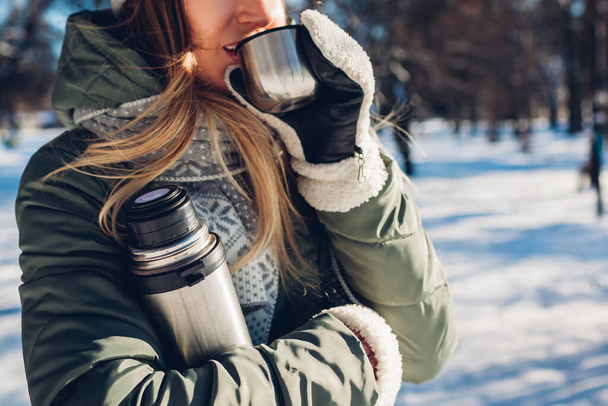 Kobieta pijąca gorącą herbatę trzymająca kolbę próżniową w parku zimowym. Napoje do ogrzania w śnieżnej mroźnej pogodzie na świeżym powietrzu podczas spaceru - Zdjęcie, obraz