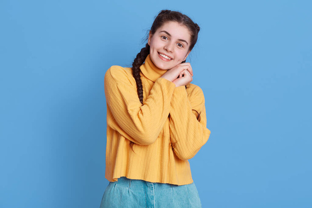Image of cute european girl with two braids wearing yellow sweater, looks smiling, looking at at camera, keeping fists together near cheeks stands against blue wall. - Fotó, kép