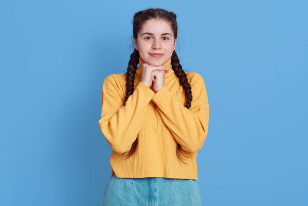 Pleasant looking Caucasian woman has charming smile and appealing look, keeps hand in fists together under chin, stands against blue wall, wearing yellow sweater and jeans. - Zdjęcie, obraz