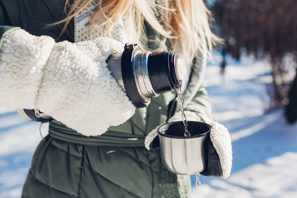 Woman pouring hot tea out of vacuum flask in winter park. Drinks to warm up in snowy frosty weather outdoors during walk - Фото, зображення
