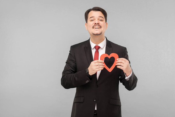 happy man holding small red heart and looking at camera with sensual look. indoor studio shot. isolated on gray background. businessman with black suit, red tie and mustache looking at camera - Φωτογραφία, εικόνα