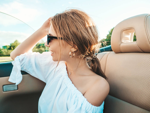 Portrait of young beautiful and smiling hipster female in convertible car. Sexy carefree woman driving cabriolet. Positive model riding and having fun in sunglasses at sunset - Foto, Bild