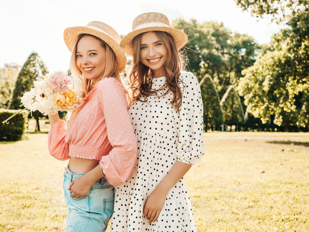 Two young beautiful smiling hipster woman in trendy summer sundress. Sexy carefree women posing in the park in hats. Positive models having fun and hugging at sunset. They holding flowers - Fotografie, Obrázek