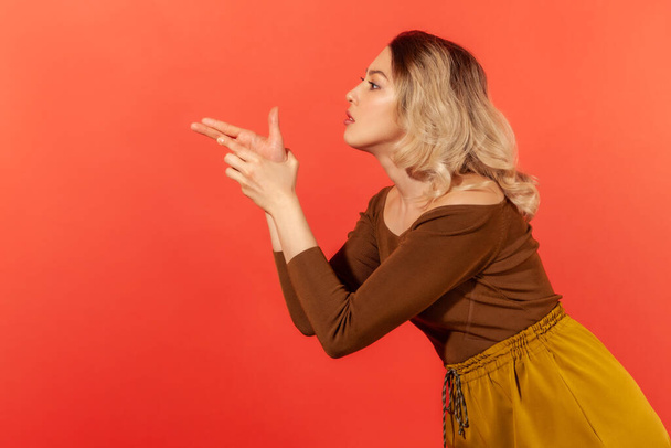 Side view portrait of dangerous criminal woman in brown blouse pointing finger gun, threatening to kill, shooting with weapon gesture right in target. Indoor studio shot isolated on red background - Photo, Image