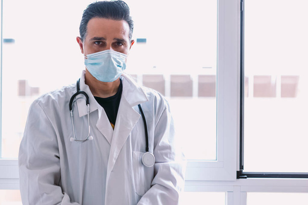 Médico de cabello negro con máscara quirúrgica, con bata blanca y estetoscopio mirando a la cámara, en una ventana. Concepto de medicina - Foto, Imagen