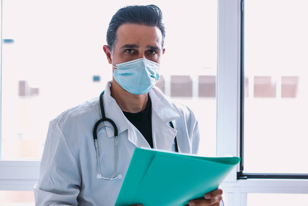 Medico con maschera chirurgica, cappotto bianco, stetoscopio e cartellina blu con un rapporto in mano davanti a una finestra. Concetto di medicina - Foto, immagini