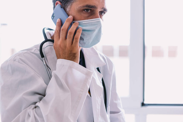 Black haired doctor in white coat and stethoscope talking on his mobile phone, at a window. Medicine concept - Foto, immagini