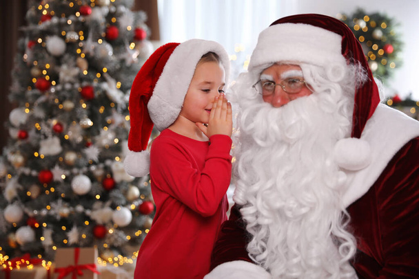 Little girl whispering in Santa Claus' ear near Christmas tree indoors - Foto, imagen