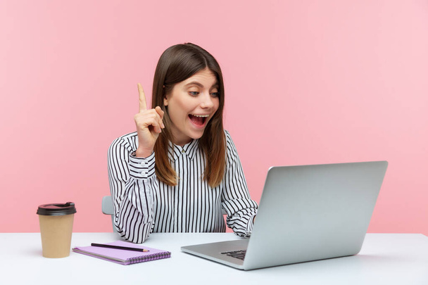 Lachende positieve vrouw kantoormedewerker in gestreept shirt wijzend vinger omhoog pratende videogesprek op laptop, geweldig idee voor het opstarten, zakelijke oplossing. Indoor studio schot geïsoleerd op roze achtergrond - Foto, afbeelding