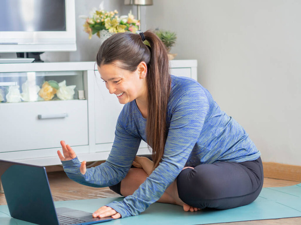 Giovane donna seduta su un tappetino yoga a casa agitando un computer portatile durante la quarantena. Ragazza sportiva con coda di cavallo in abbigliamento sportivo iniziare un allenamento a casa. - Foto, immagini