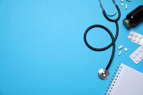 Stethoscope, pills and notebook on light blue background, flat lay. Space for text - 写真・画像