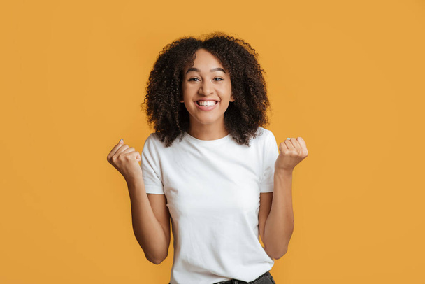 Gagner souriant jeune femme afro-américaine avec les cheveux bouclés lever les poings et se réjouir de la victoire en ligne - Photo, image