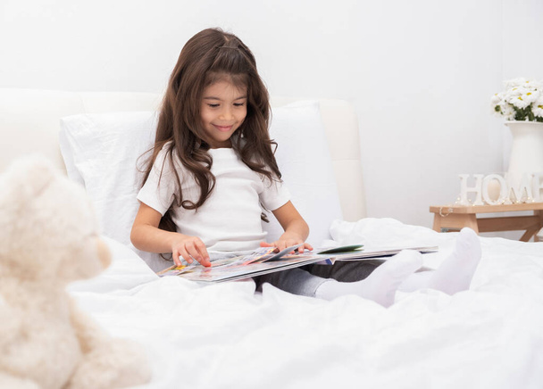 щаслива маленька брюнетка дівчинка сидить на ліжку вдома читаючи книгу
. - Фото, зображення
