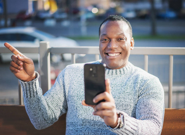Stylish happy young african american man in a gray sweater sitting on a bench and talking in a smartphone - Foto, immagini