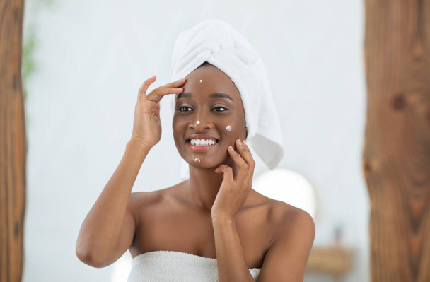 Happy african american lady with white towel on head looks in mirror and smears face with cream - Foto, Imagem