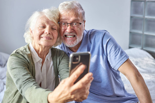 caucasian cheerful joyful senior couple sitting on a couch taking photo on smartphone - Zdjęcie, obraz