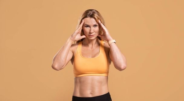 Surullinen lihaksikas keski-ikäinen nainen urheiluvaatteissa painaa käsistä päähän kärsii päänsärystä - Valokuva, kuva