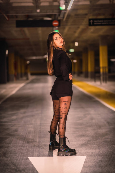 Young Caucasian model perched on the asphalt of an empty underground car park. Night urban session in the city - Foto, Imagem