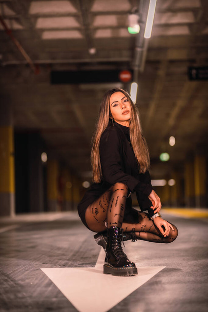 Young caucasian brunette in black jacket on the white arrow of the ground in an empty underground car park. Night urban session in the city - Photo, image