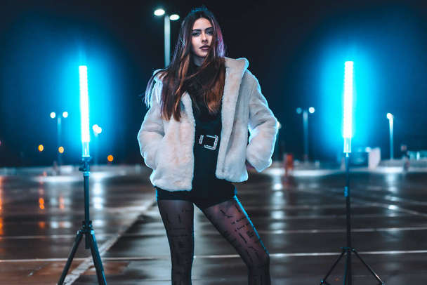 Young Caucasian brunette with pink wool jacket in an empty car park illuminated with blue light. Night urban session in the city - Foto, Imagem