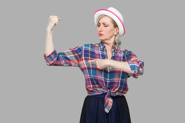 Strong and independent alone. Portrait of proud modern stylish mature woman in casual style with white hat standing and pointing finger at her biceps. indoor studio shot isolated on gray background. - 写真・画像