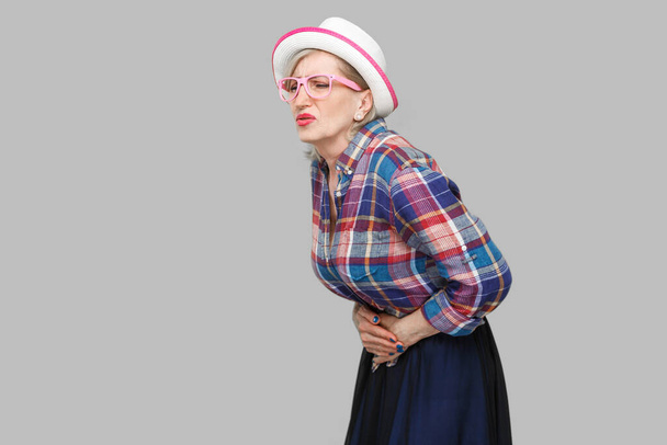 Maagpijn. Profiel zijaanzicht portret van moderne stijlvolle volwassen vrouw in casual stijl met hoed en bril staan en houden haar pijnlijke buik. indoor studio schot geïsoleerd op grijze achtergrond - Foto, afbeelding