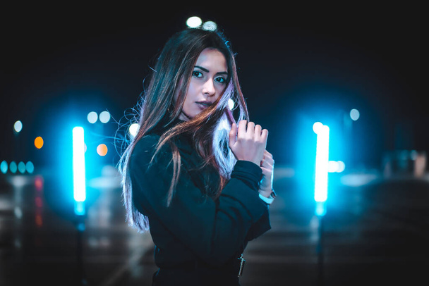 Pretty Caucasian brunette in a pink wool jacket in an empty car park illuminated with blue light. Night urban session in the city - Фото, зображення
