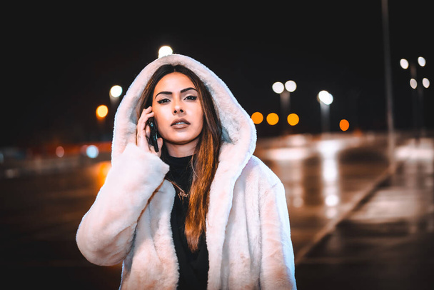 Caucasian brunette talking on the phone in a pink wool jacket in an empty car park. Night urban session in the city - Zdjęcie, obraz