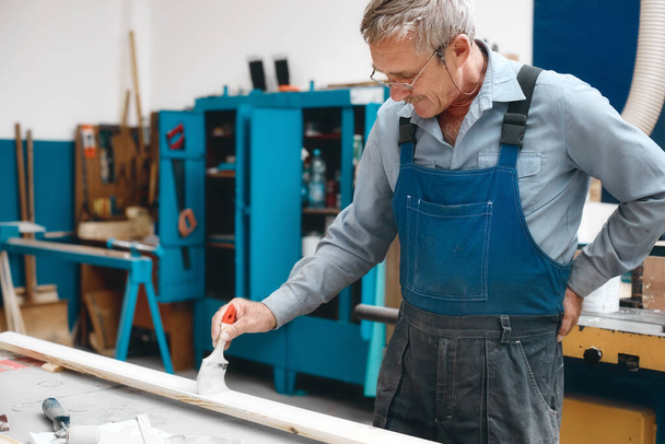 Um fabricante de armários idoso em macacões e óculos pinta uma tábua de madeira com um pincel em uma bancada de trabalho em uma loja de carpintaria.  - Foto, Imagem