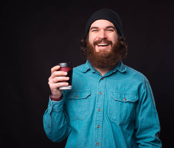 Boldog szakállas férfi kezében egy csésze kávét, hogy menjen át a sötét háttér - Fotó, kép