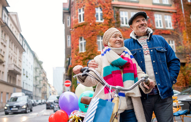 Happy senior couple with bicycle and balloons outdoors on street in city. - Fotografie, Obrázek
