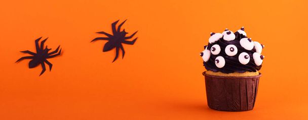 black cupcake with lots of marshmallow eyes for halloween on an orange background with black spiders. mystical scary background - Photo, Image