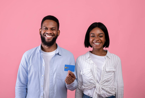 Portrait of happy black millennial couple promoting bank services, showing credit card on pink studio background - Photo, Image