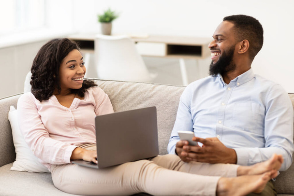 Couple afro-américain assis sur le canapé, en utilisant un ordinateur portable et un smartphone - Photo, image