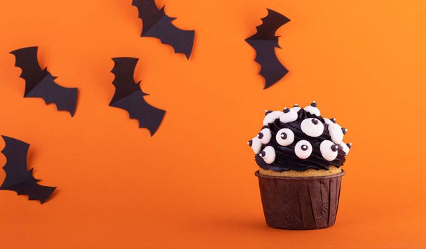 black cupcake with lots of marshmallow eyes for halloween on an orange background with black spiders. mystical scary background - 写真・画像