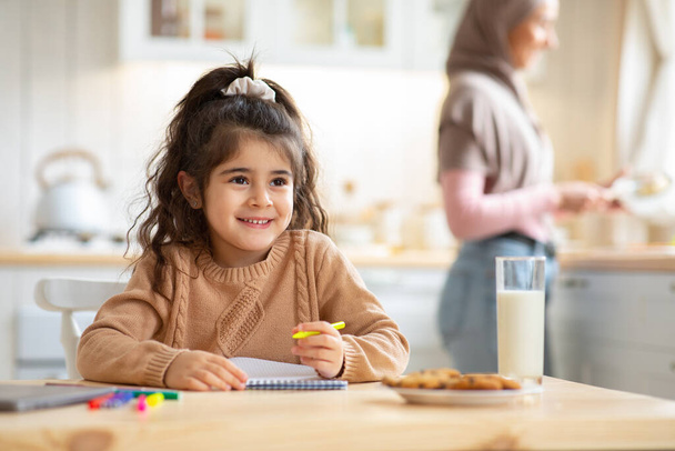 bonito menina desenho no cozinha enquanto ela muçulmano mãe cozinhar almoço - Foto, Imagem