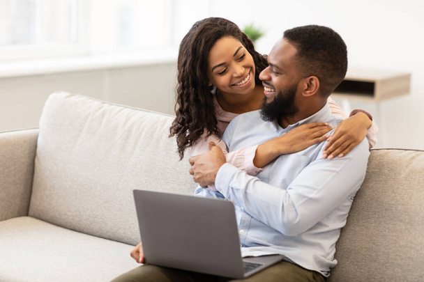 Feliz pareja afro-americana amorosa sentada en el sofá, usando el ordenador portátil - Foto, imagen
