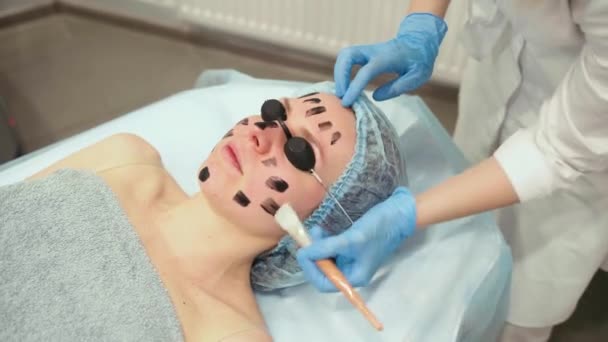 Δερματολόγος μουτζουρώνει μαύρη μάσκα στο πρόσωπο για φωτορεπορτάζ λέιζερ και ξεφλούδισμα άνθρακα. Δερματολογία και κοσμετολογία. Χρήση χειρουργικού λέιζερ. - Πλάνα, βίντεο